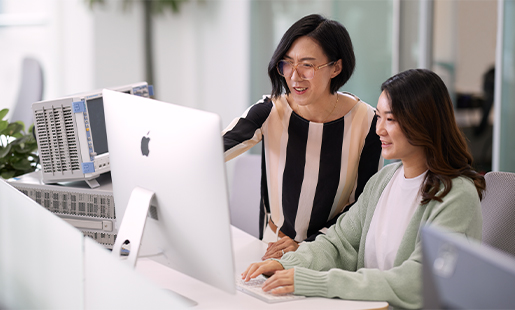Apple 디스플레이 앞에 앉아 있는 동료 옆에 서 있는 Ella.