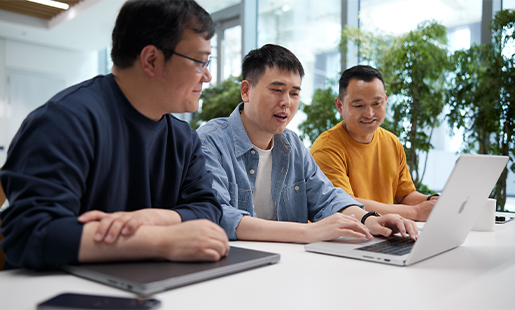 밝은 창과 식물들이 있는 공간에서 두 동료 사이에 앉아 MacBook으로 타자를 치고 있는 Ken.