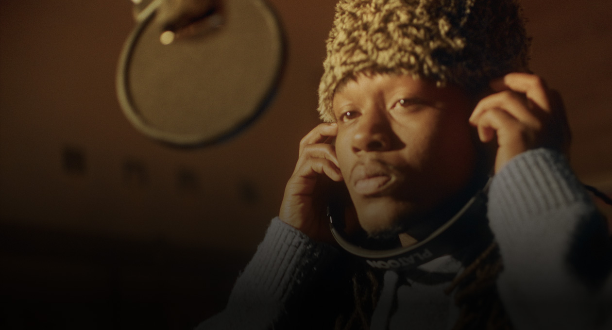 Photo de Kiddo K dans un studio de musique; il se tient derrière un micro d’enregistrement et porte un casque d’écoute.
