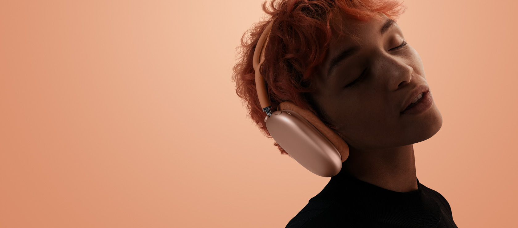 Une personne, la tête inclinée et les yeux fermés, porte des AirPods Max 2, couleur orange, sur un fond rose orangé