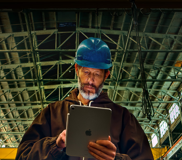 iPad, argent, avec un Apple Pencil fixé sur le côté, tenu par une personne portant une veste brune et un casque de protection bleu dans un local industriel avec des poutres de métal au plafond