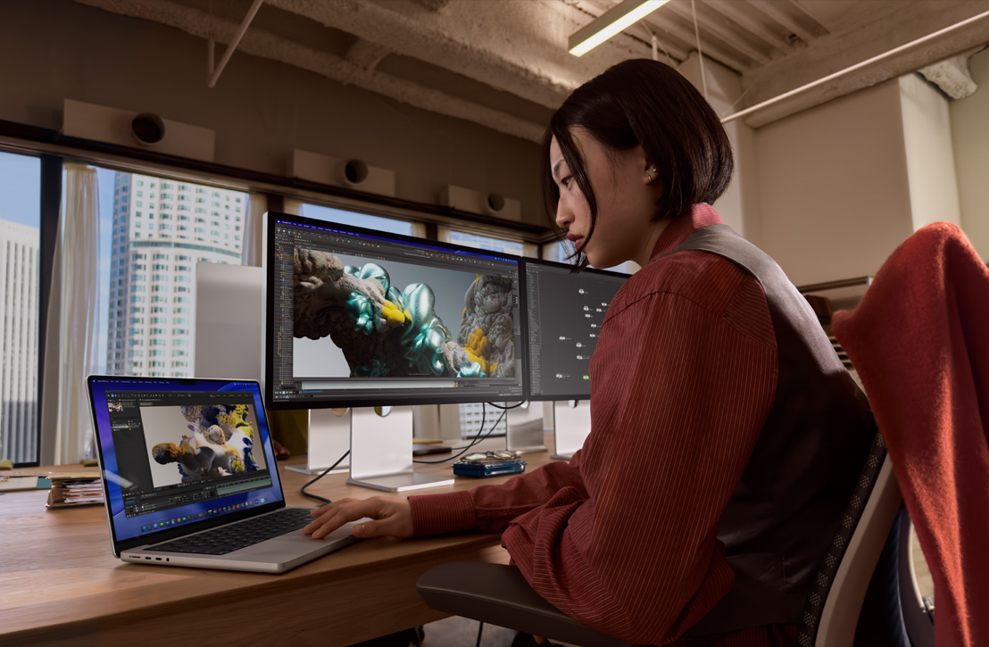 Une personne travaille à son bureau sur un MacBook Pro branché à un moniteur externe; les deux écrans affichent un rendu 3D; des immeubles sont visibles par la fenêtre