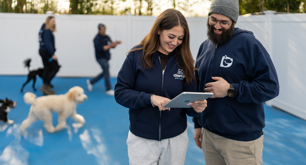 Deux personnes portant des chandails à capuchon aux couleurs de leur entreprise regardent un iPad ensemble dans une garderie pour chiens, des chiens et du personnel sont visibles en arrière-plan