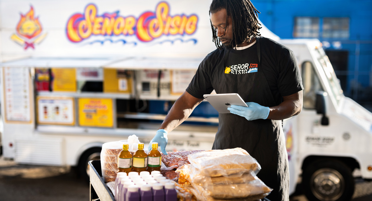 Une personne portant un t-shirt aux couleurs de son entreprise et des gants consulte un iPad tout en préparant de la nourriture à une table, un camion-restaurant est en arrière-plan