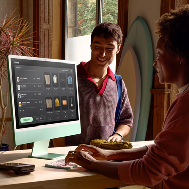En medarbetare sitter bakom disken i en butik som säljer surfbrädor och andra föremål och använder en iMac för att slutföra ett köp åt en kund på andra sidan disken.