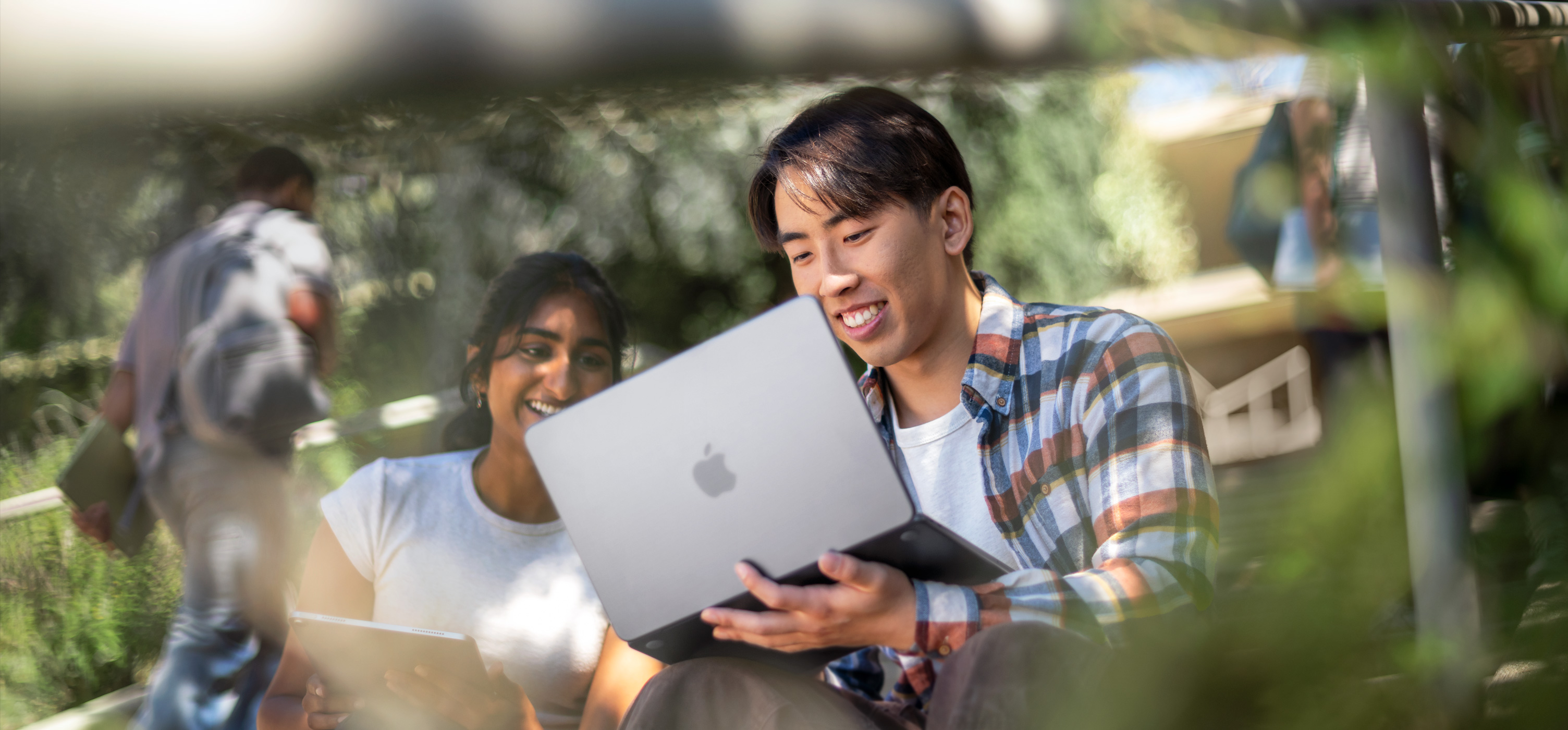 兩名面帶微笑的學生在戶外使用 MacBook 合力完成作業，MacBook 放在其中一人腿上