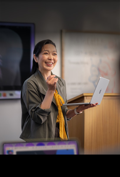 教授在課堂上拿著銀色 MacBook 對學生做簡報，展示 MacBook 卓越的效能和持久的電池續航力