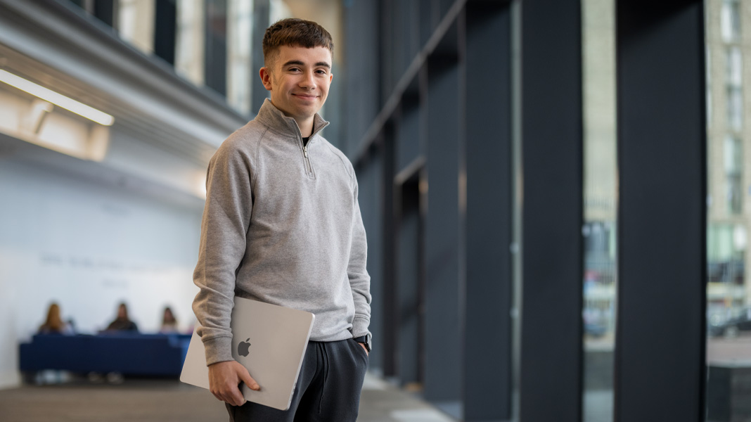 Uno studente in piedi nel corridoio di un edificio moderno tiene in mano un portatile MacBook e sorride alla fotocamera
