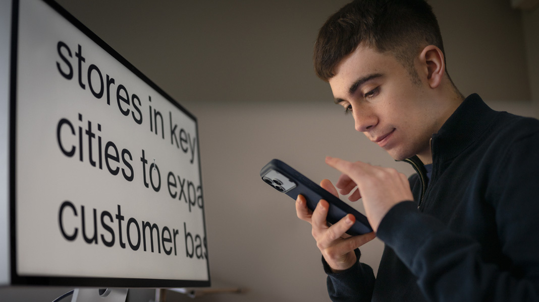 Uno studente usa un iPhone mentre è seduto davanti a un monitor che mostra i contenuti del corso di economia con il testo ingrandito