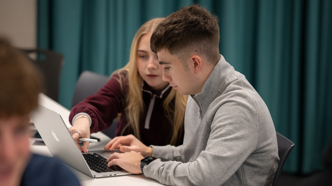Uno studente e una studentessa lavorano insieme su un MacBook, lei indica lo schermo mentre lui digita