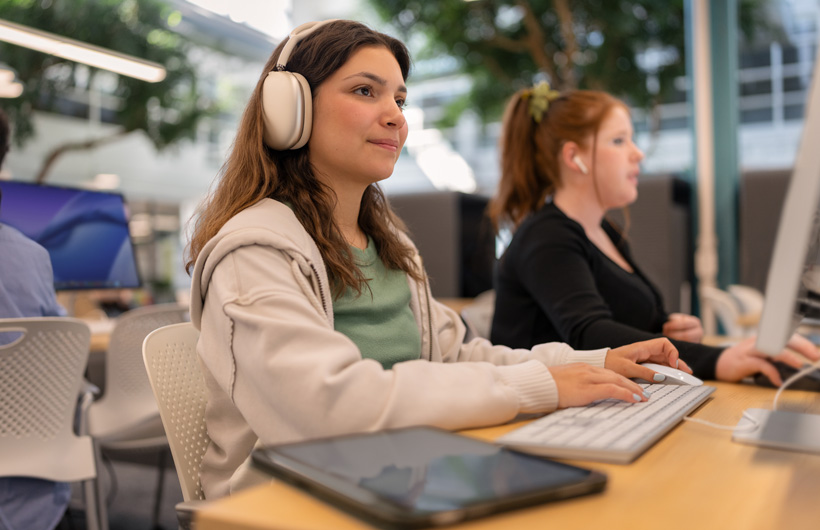 Una estudiante con unos AirPods Max sentada frente a una computadora escribe en el teclado, con una tablet cerca, mientras otra estudiante trabaja sentada junto a ella.