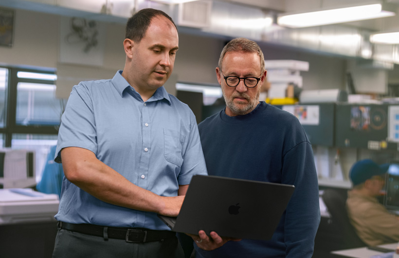 Dos hombres en una estación de trabajo miran juntos una MacBook, uno la sostiene mientras el otro señala la pantalla.