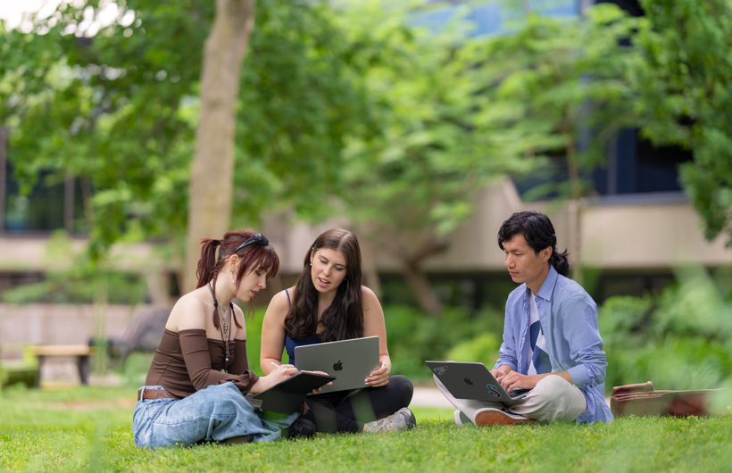 Tres estudiantes sentados en el césped en un campus al aire libre, una usa una MacBook y otro observa.