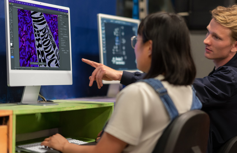 Dos estudiantes sentados frente a dos dispositivos iMac que muestran modelos coloridos, uno de los estudiantes señala la pantalla.