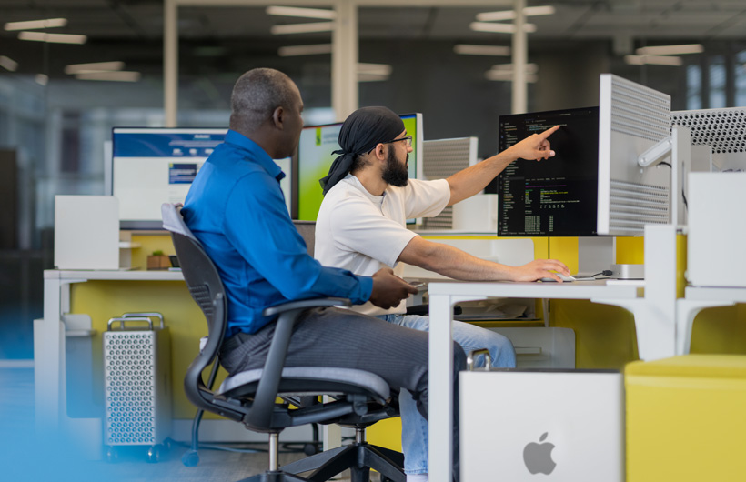 Dos estudiantes sentados frente a varios dispositivos iMac, uno de ellos señala una pantalla.