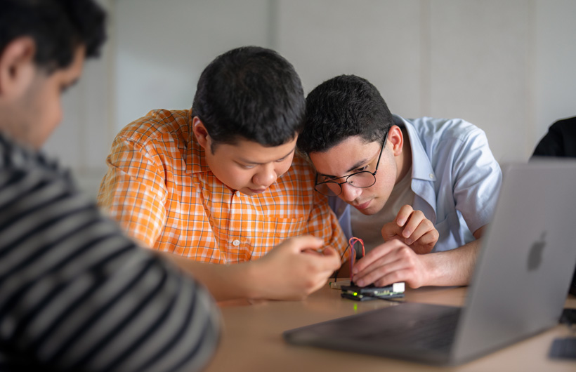 Dos estudiantes examinan de cerca una placa de circuito junto a una MacBook, mientras otra persona los observa.