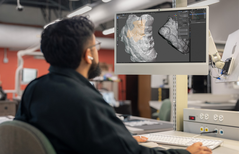 Un estudiante sentado en un escritorio revisa dos modelos 3D que se muestran uno al lado del otro en monitores de iMac.