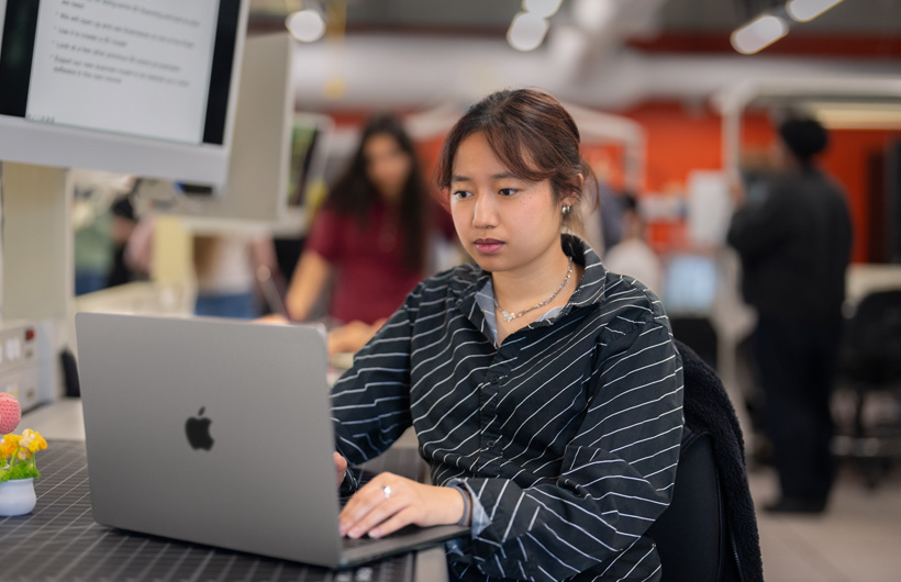 Una estudiante sentada en un escritorio en un salón de clases moderno usa una MacBook.