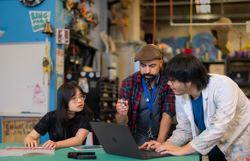 Tres estudiantes reunidos alrededor de una MacBook ubicada en la mesa de un colorido taller repleto de herramientas y obras de arte.