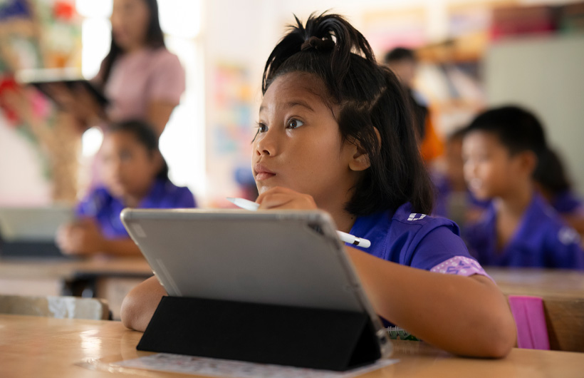 Una niña en su pupitre voltea con atención al frente de la clase. Usa un iPad con un Apple Pencil.