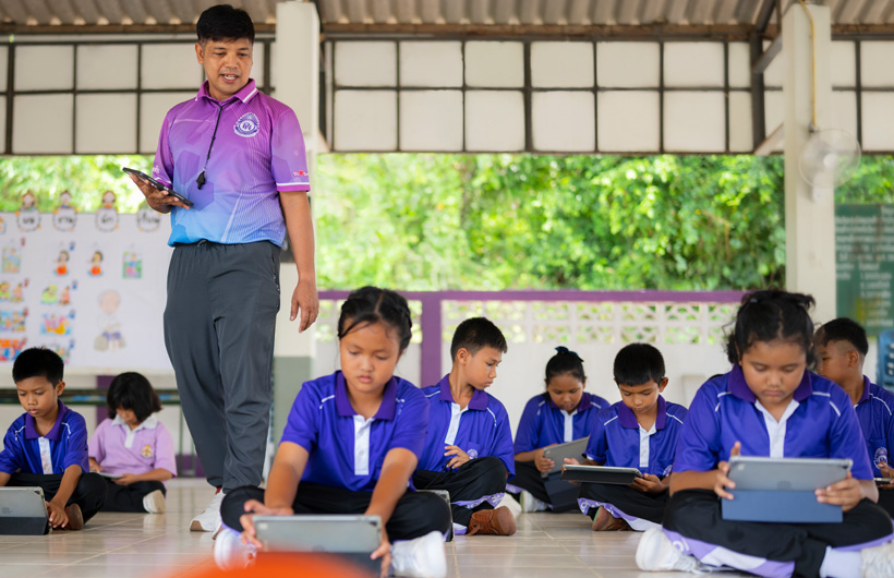En un salón de clases al aire libre, un profesor de educación física camina entre los estudiantes, que están sentados en el piso con sus iPad en la mano.