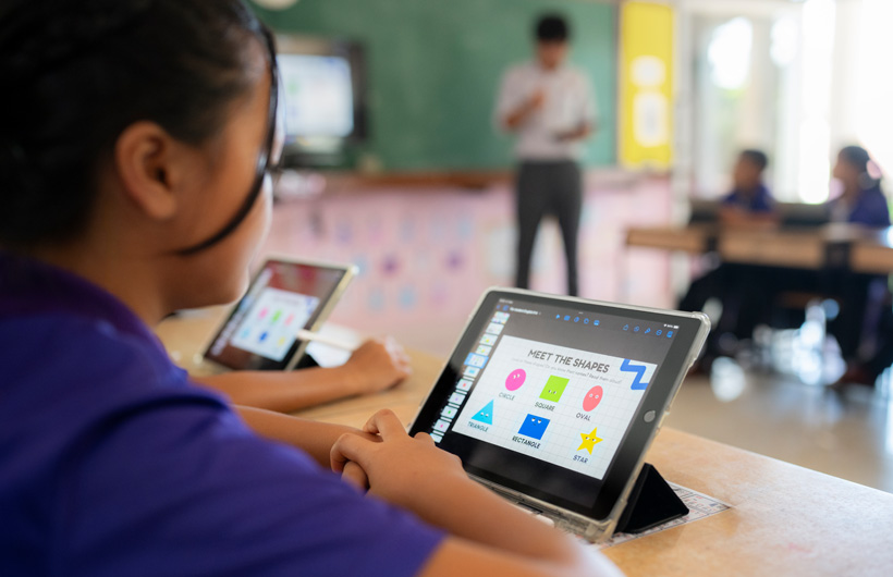 Primer plano de una estudiante en su pupitre con un iPad. En la pantalla hay unas figuras de colores con el título “Conoce las formas”.