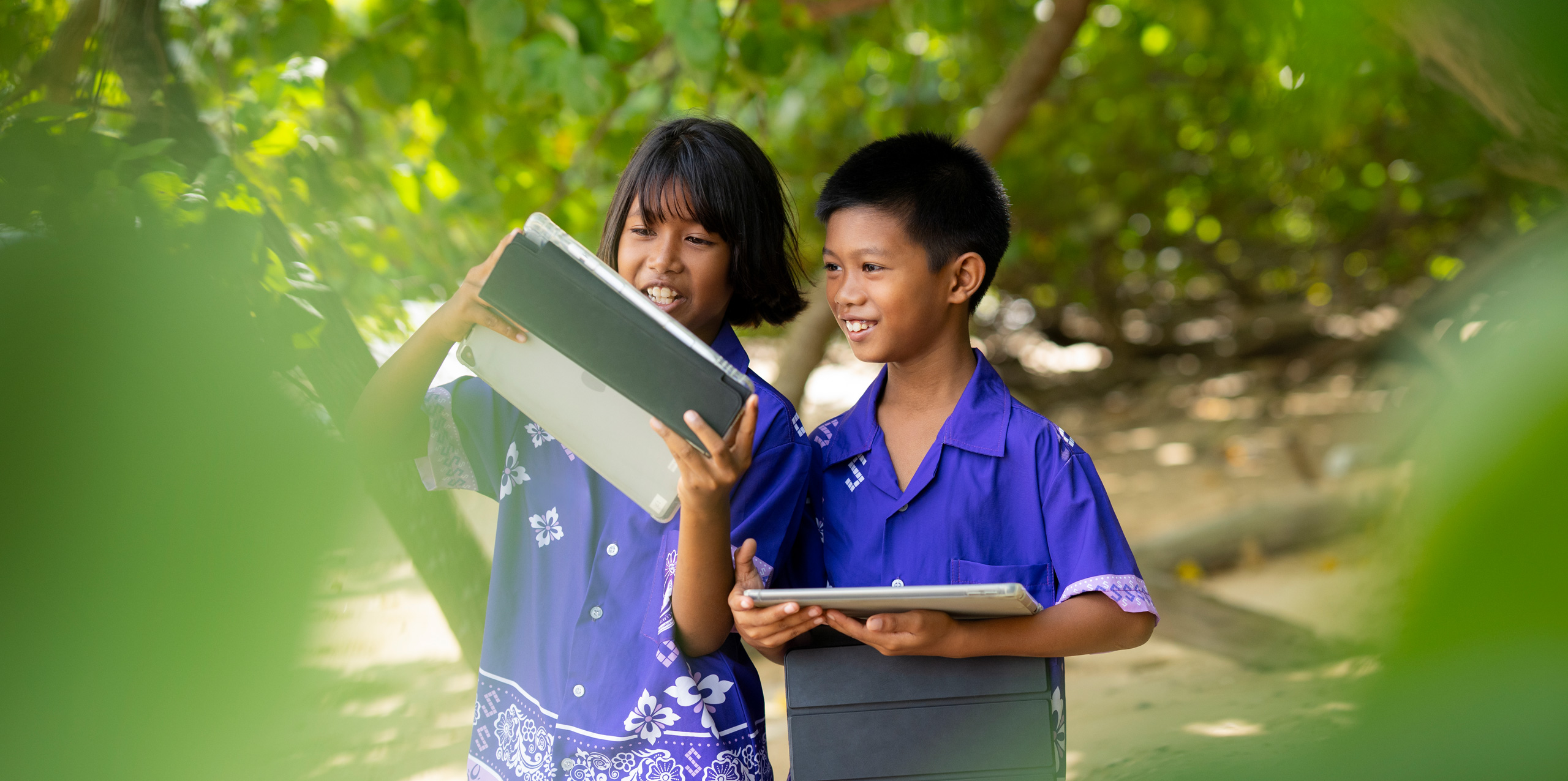 Dos estudiantes de primaria, un niño y una niña, de pie al aire libre entre árboles tropicales, cada uno con un iPad en la mano. Ambos sonríen y ven atentos la pantalla del iPad de la niña.