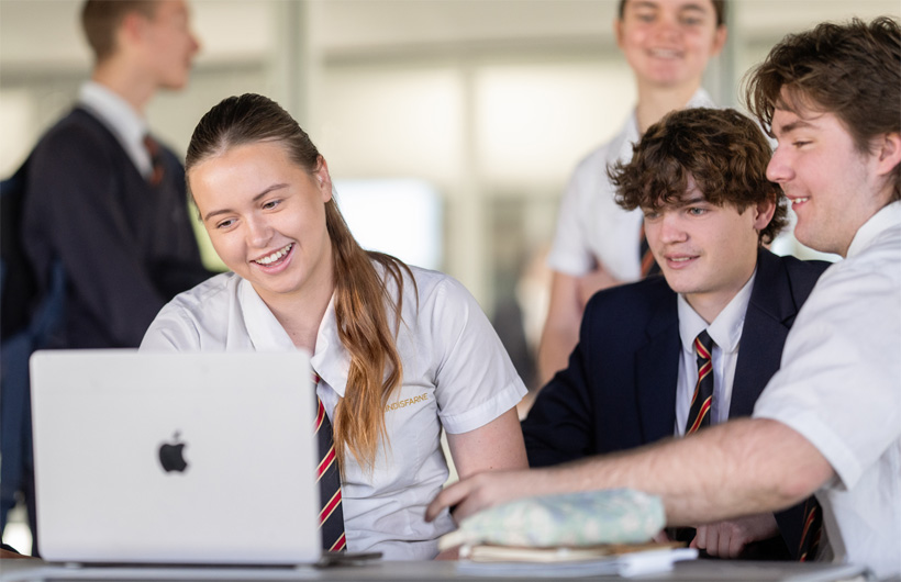 Un grupo de estudiantes con uniforme escolar se reúne alrededor de una MacBook, sonriendo y colaborando.