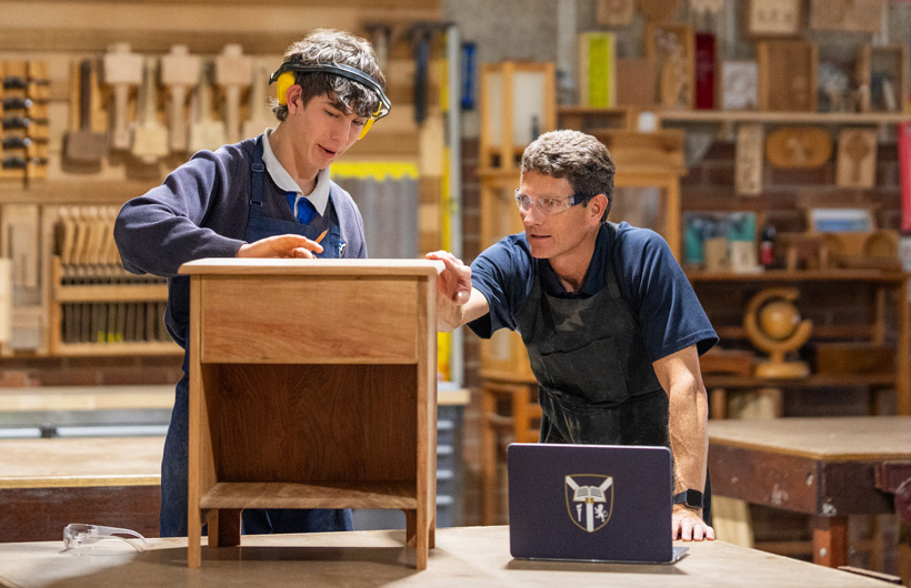 Un profesor de carpintería y un estudiante examinan un pequeño mueble de madera. Hay una laptop de la escuela en la mesa de trabajo delante de ellos.