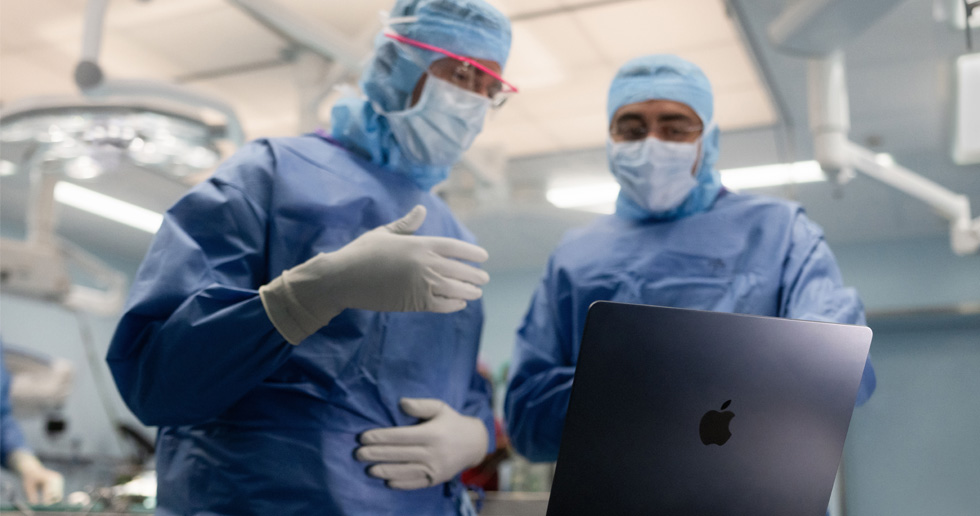 Dos cirujanos en el quirófano miran un MacBook, y uno hace gestos con la mano mientras el otro observa