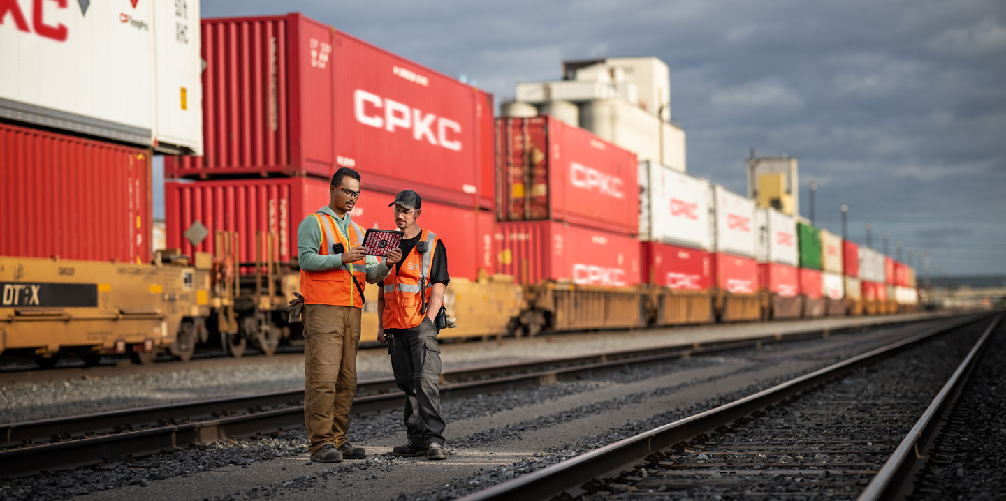 Dos trabajadores del ferrocarril consultan un iPad de pie en un deposito ferroviario junto a un tren parado que transporta contenedores