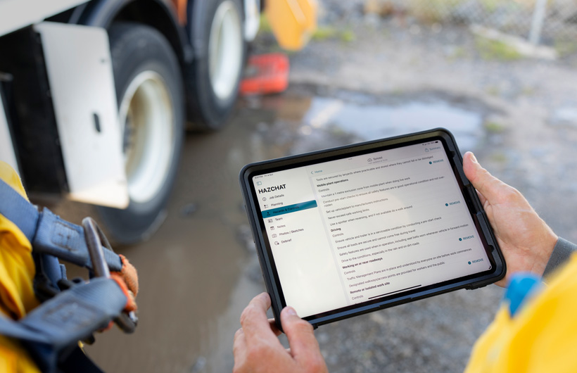 Mãos seguram um iPad com o app HazChat aberto, que exibe detalhes do trabalho, planejamento, riscos e controles, e uma pessoa está em pé no local de trabalho ao lado de uma poça de lama