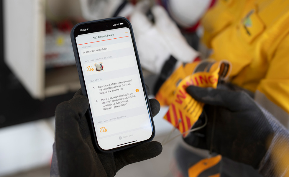 Mão segura um iPhone que exibe instruções para realizar um teste no quadro de distribuição, com as palavras 13C Process Step 3 na tela
