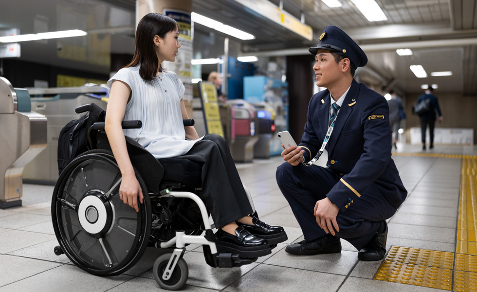 オフィススタイルに身を包んだ車椅子の女性と駅係員がホームで会話をしている