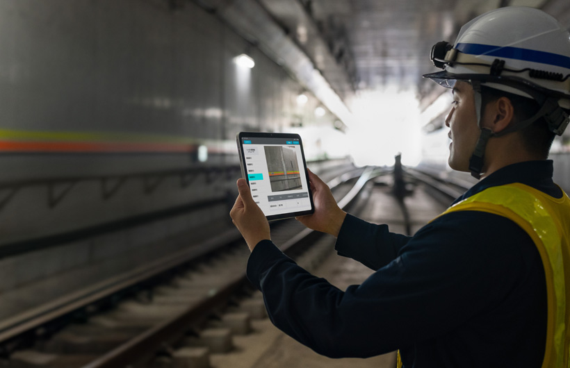 Un addetto alla manutenzione delle gallerie di Tokyo Metro che tiene in mano un iPad per acquisire un’immagine dei binari mentre il suo collega guarda.