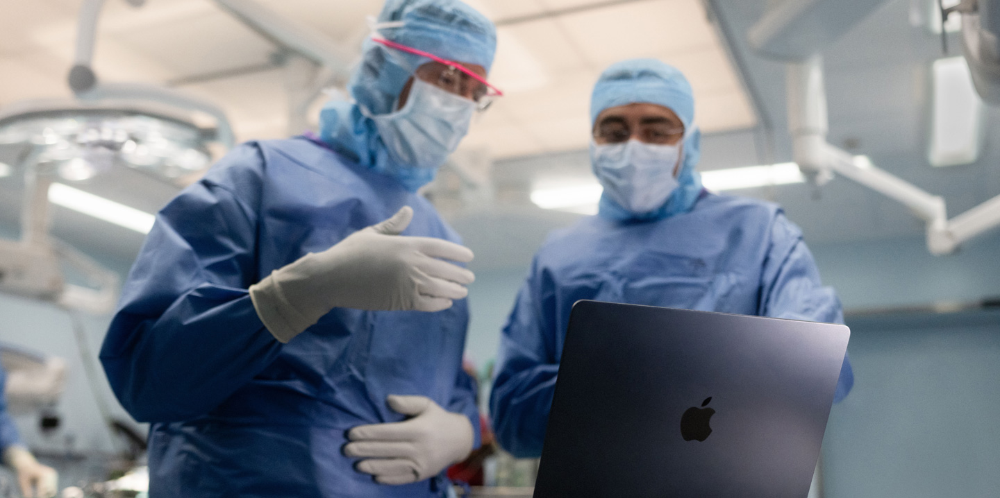 수술실에 있는 두 명의 외과의가 MacBook을 보며, 한 명은 손으로 손짓을 하고 다른 한 명은 지켜보고 있는 모습