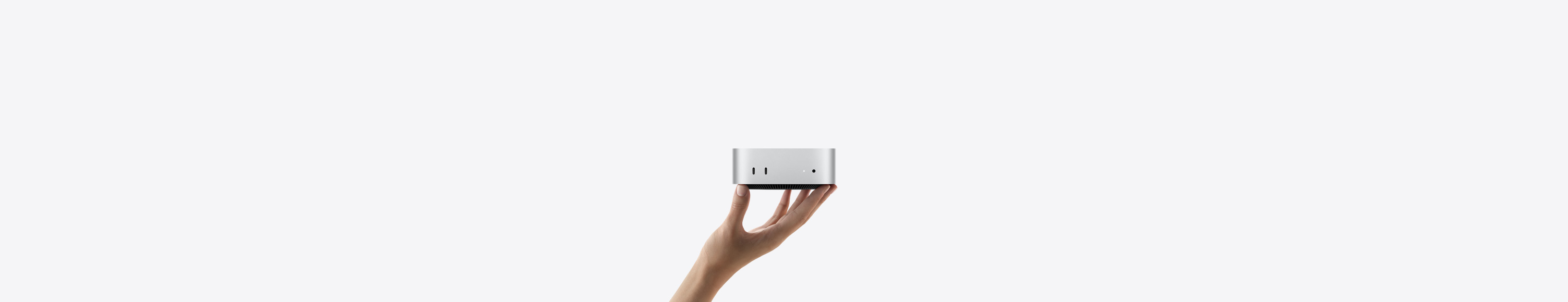 Front view of Mac mini balanced on the fingertips of an up-stretched hand, front shows two Thunderbolt ports, status indicator light and headphone jack, tapered black base at bottom, flat top, rounded sides, straight edges, silver colour