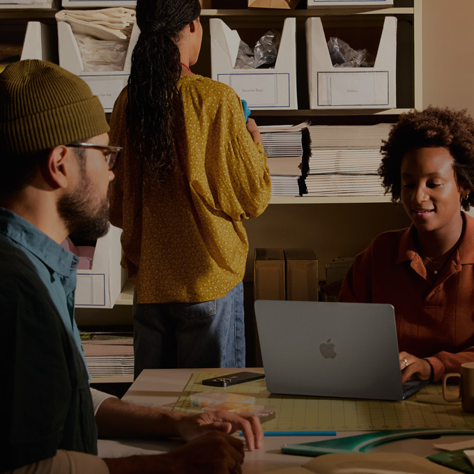 Trois personnes dans un bureau, deux d’entre elles sont assises et semblent consulter du contenu à l’écran d’un MacBook, tandis que la troisième fait face à un tableau affichant plusieurs graphiques